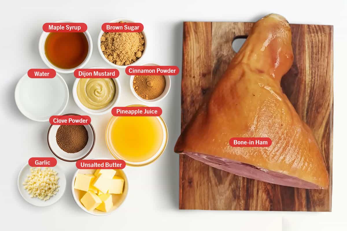 Top shot of the following ingredients: bone-in ham, maple syrup, brown sugar, water, Dijon mustard, cinnamon powder, clove powder, pineapple juice, garlic, and unsalted butter on a white surface.