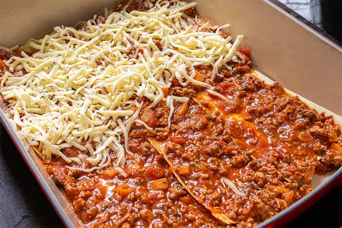 Image of lasagna being assembled with layers of pasta sheets, rich ragu sauce, and shredded mozzarella cheese.