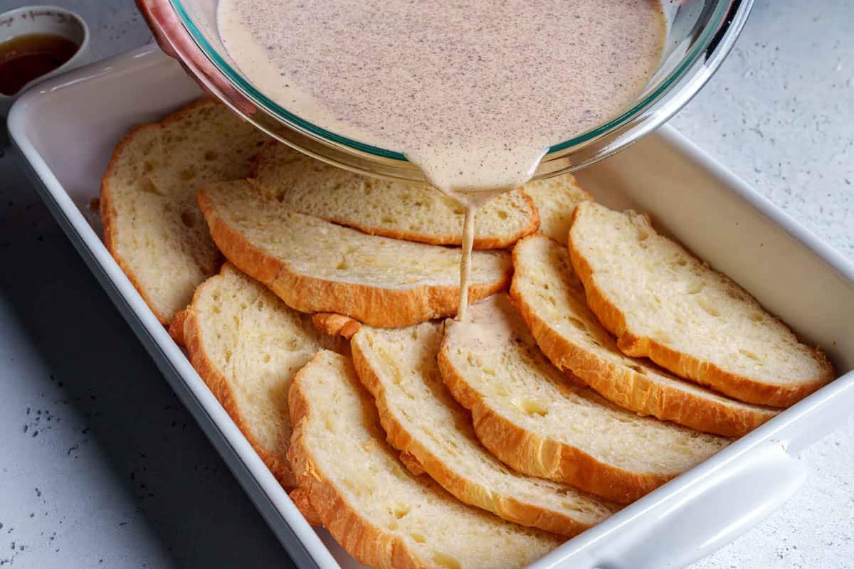 Half of the egg mixture is being poured over the halved croissants in a greased pan.