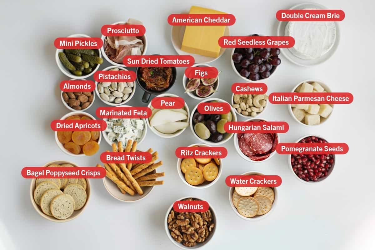 Top shot of ingredients: Mini pickles, Prosciutto, american cheddar, red seedless grapes, double cream brie, almonds, pistachios, sun dried tomatoes, figs, cashews, italian parmesan cheese, dried apricots, marinated feta, pear, olives, hungarian salami, pomegranate seeds, bagel poppyseed crisps, bread twists, ritz crackers, water crackers, and walnuts.