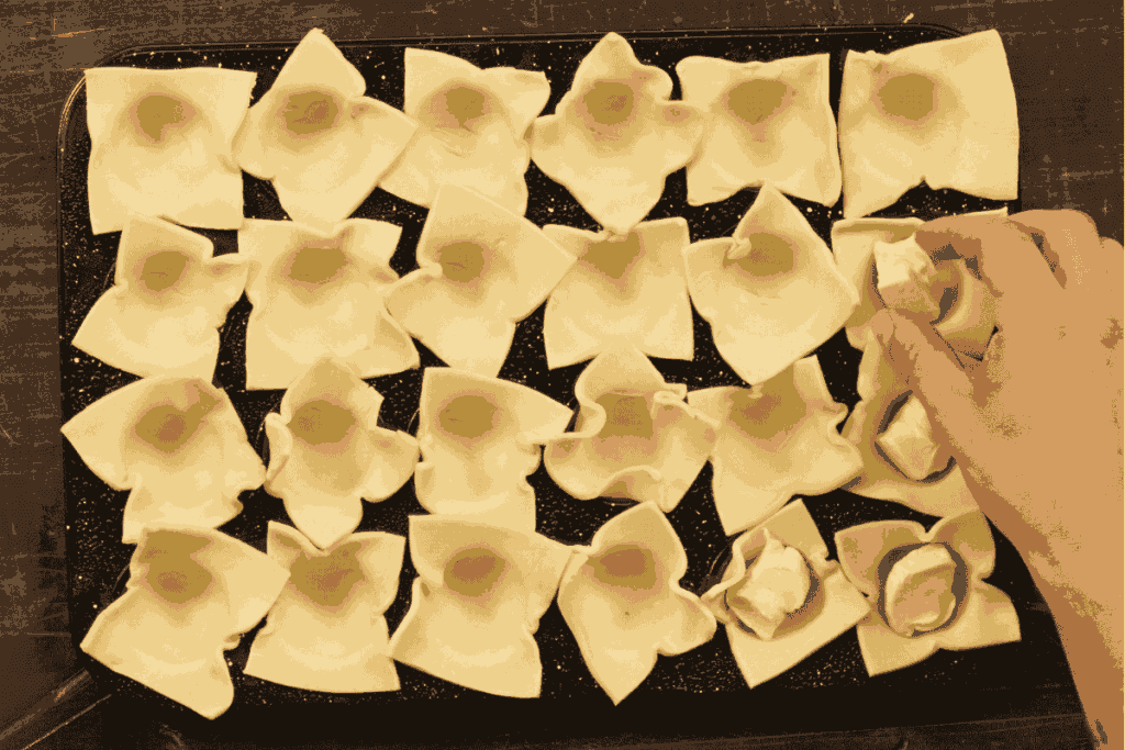 a top down picture of a 24 hole mini muffin tray filled with puff pastry and a hand placing a brie cheese in each hole