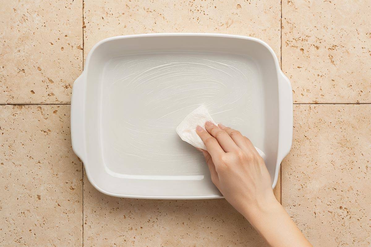 Overhead shot of a white ceramic baking dish being lightly greased with a paper towel.