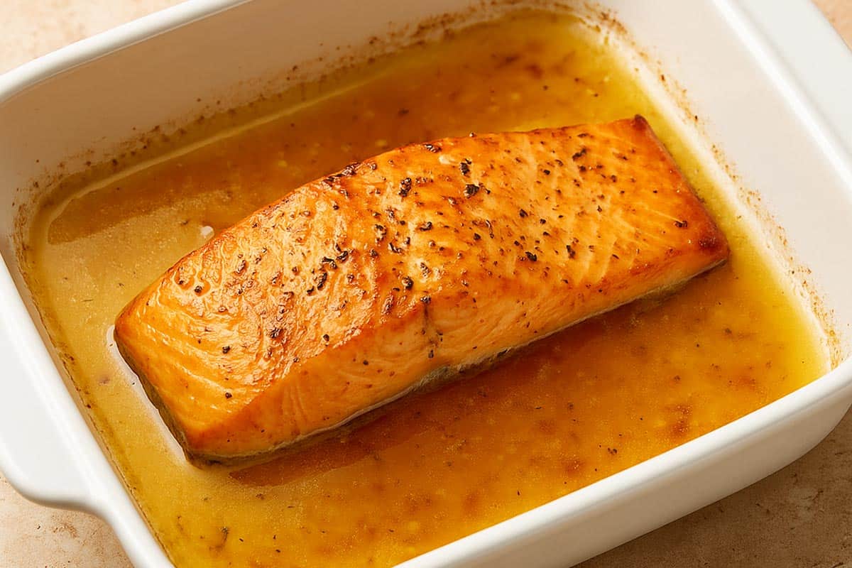 Baked salmon fillet in a white ceramic dish, resting in a pool of golden lemon butter sauce.