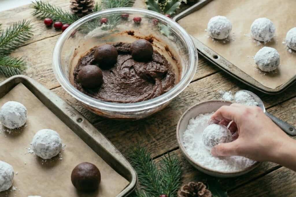 a side view of a hand in icing sugar bowl rolling a ball of brown mix in it forming a cookie dough covered in sugar. There are two paper lined baking racks next to a bowl of cookie dough.