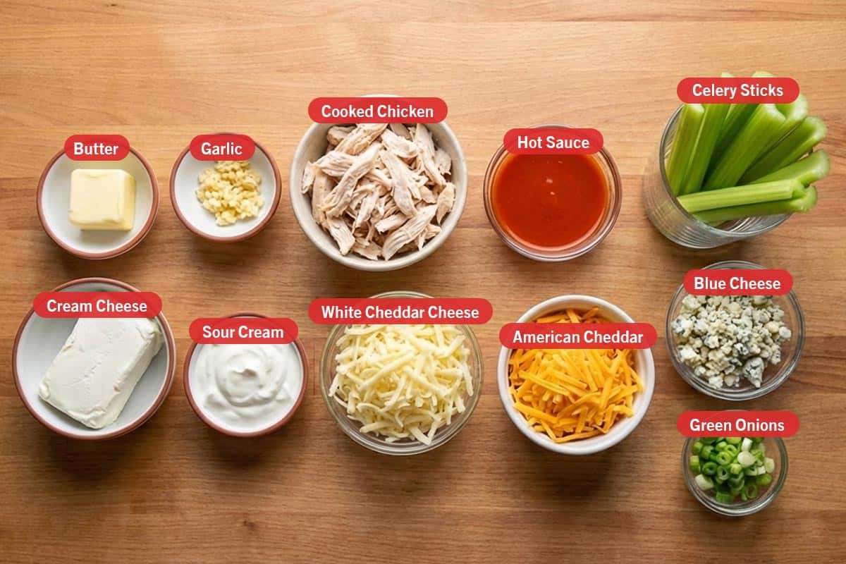 Top shot of ingredients: butter, garlic, cooked chicken, hot sauce, celery sticks, cream cheese, sour cream, white cheddar cheese, american cheddar, blue cheese, and green onions.