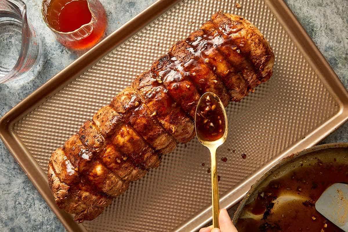 a long pork loin resting on a perforated oven rack ready for baking with a spoon of glaze covering the roast