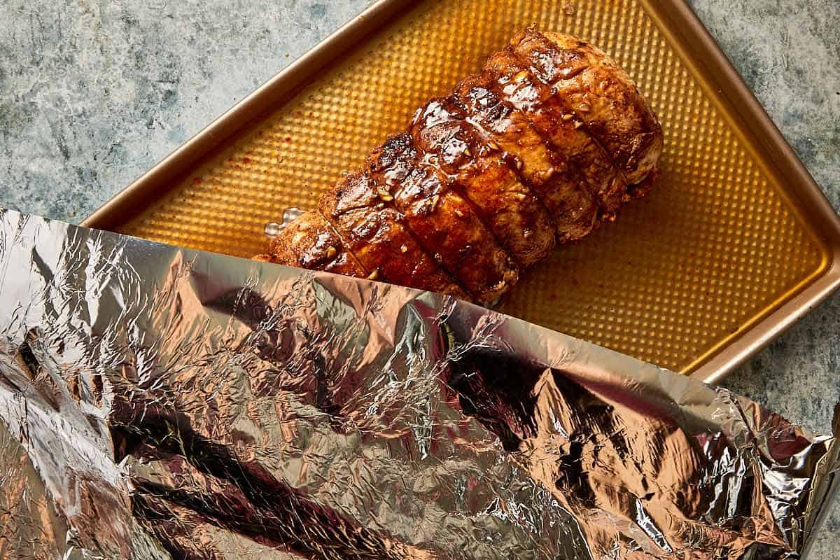 a long pork loin resting on a perforated oven rack ready for baking with a spoon of glaze covering the roast and aluminum foil being placed on top of the rack to cover the roast