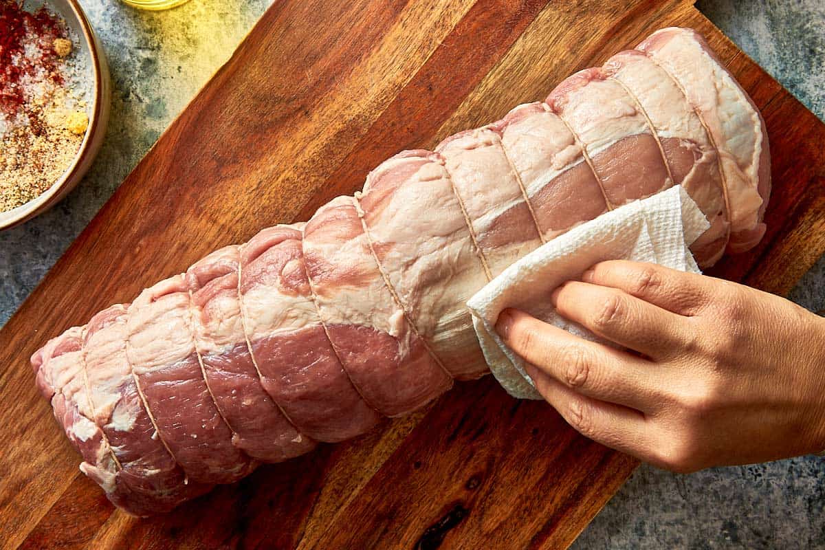 A top down view of a long pork loin chop on a board being patted down by a paper towel