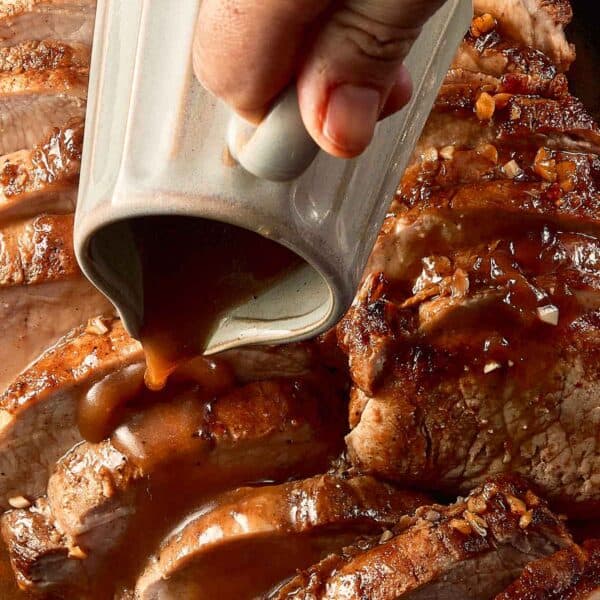 top down view of pork loin roast cut into half inch pieces in a log formation. fully cooked on the inside covered with gravy with a jug pouring the gravy on top