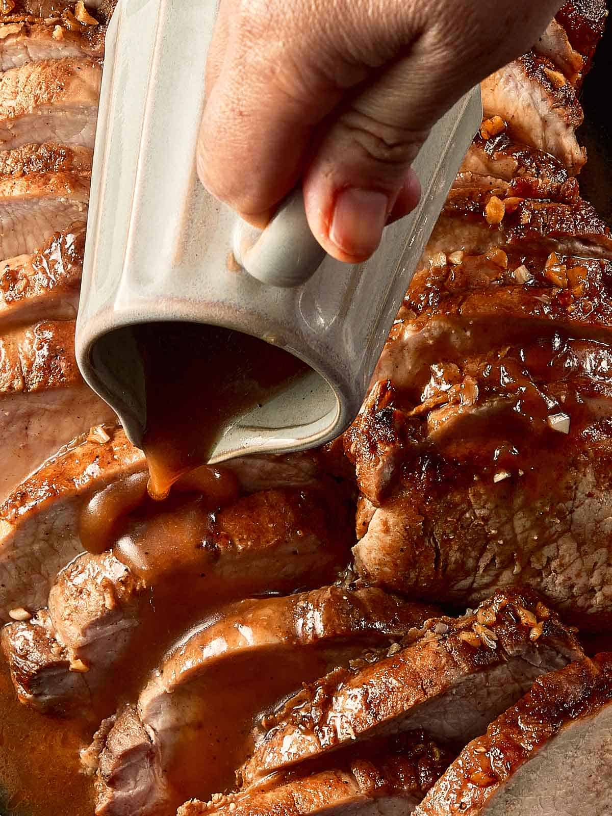top down view of pork loin roast cut into half inch pieces in a log formation. fully cooked on the inside covered with gravy with a jug pouring the gravy on top