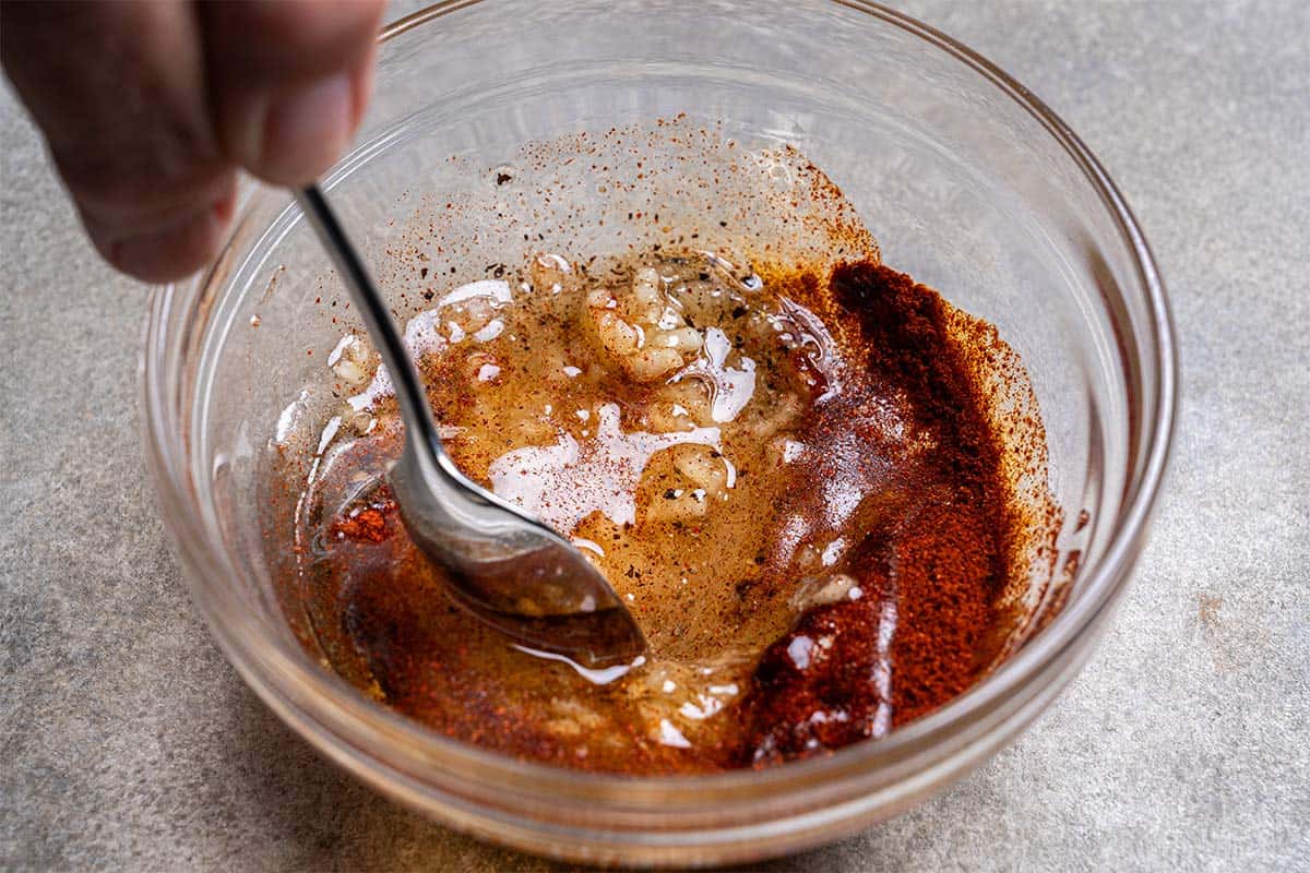 Oil, garlic, onion powder, salt, paprika, and pepper are being combined in a clear bowl with a spoon.