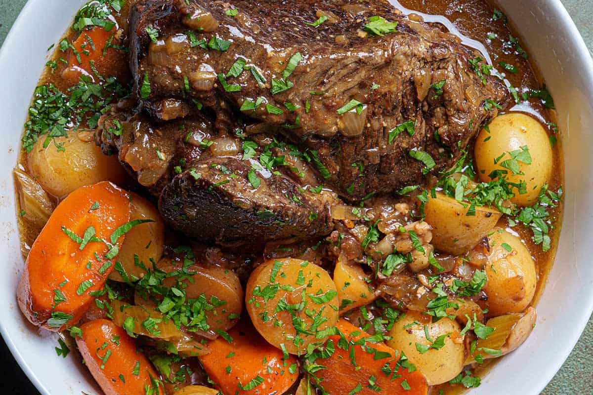 An image of the finished pot roast served on a round white plate, accompanied by potatoes and carrots, and sprinkled with fresh parsley.