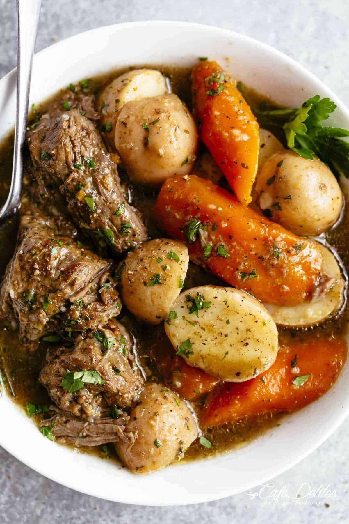 An image of a perfectly cooked pot roast, soaked in jus, is ready to be served in a white bowl, accompanied by potatoes and carrots, and garnished with parsley.