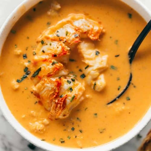 Cooked lobster flesh submerged in an orange soup in a white bowl. A black spoon is scooping some of the orange liquid in a bowl and spoon.