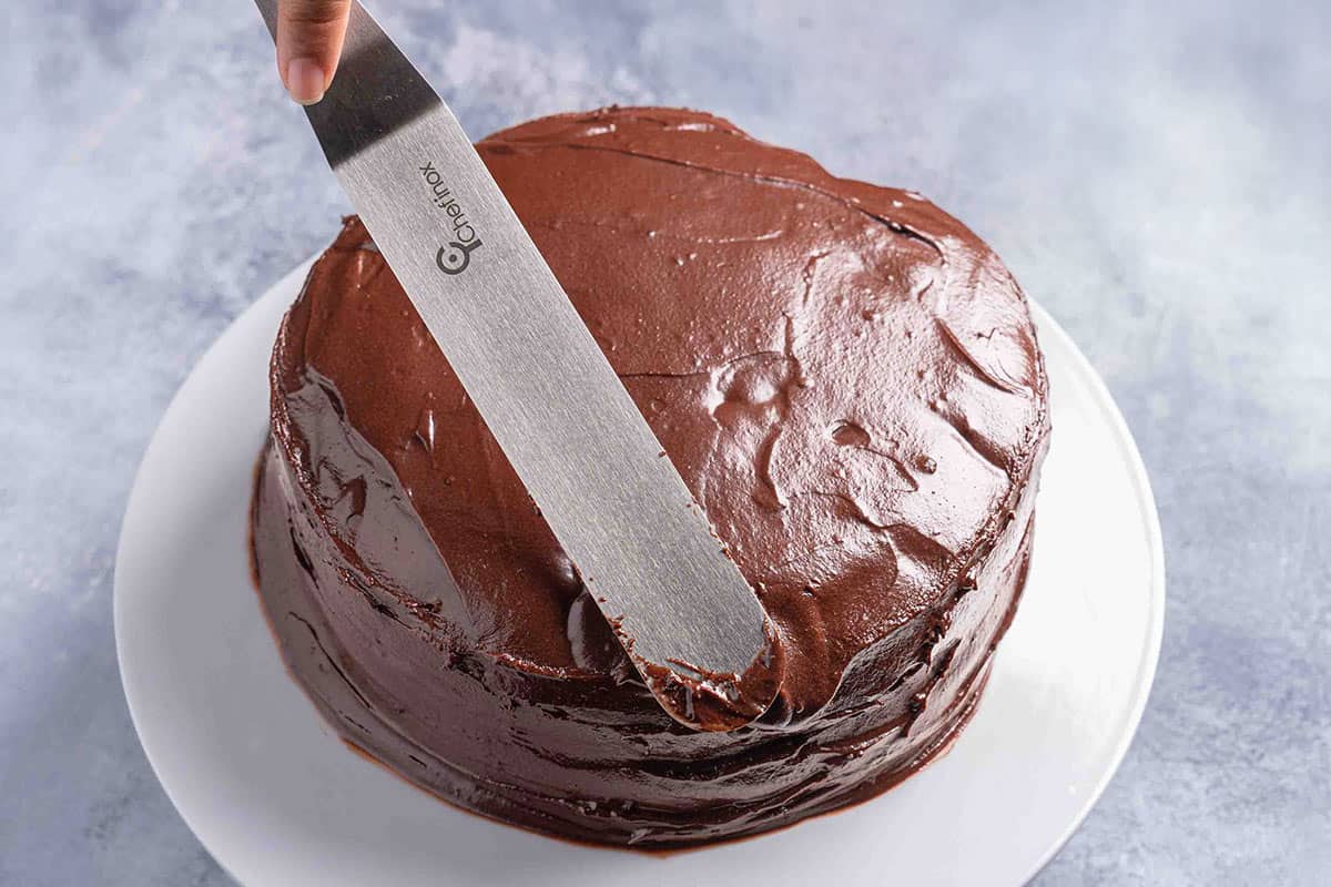 frosted chocolate cake sitting on a cake stand with a spatula frosting 