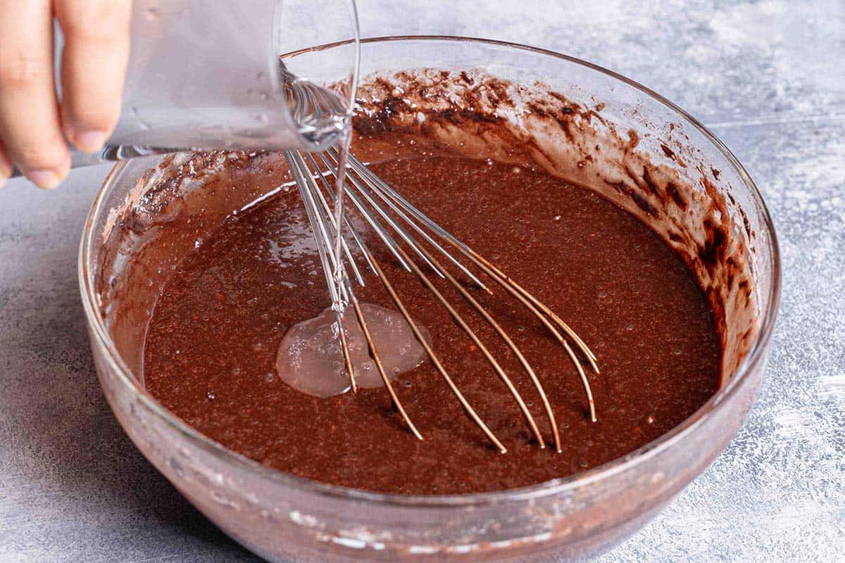 boiling water being poured into the brown chocolate cake mix in a clear bowl