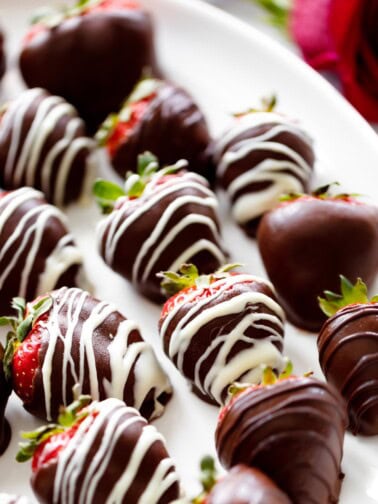 A white plate full of alcohol infused chocolate coated 
strawberries. A rose sits behind the plate for romantic decoration.