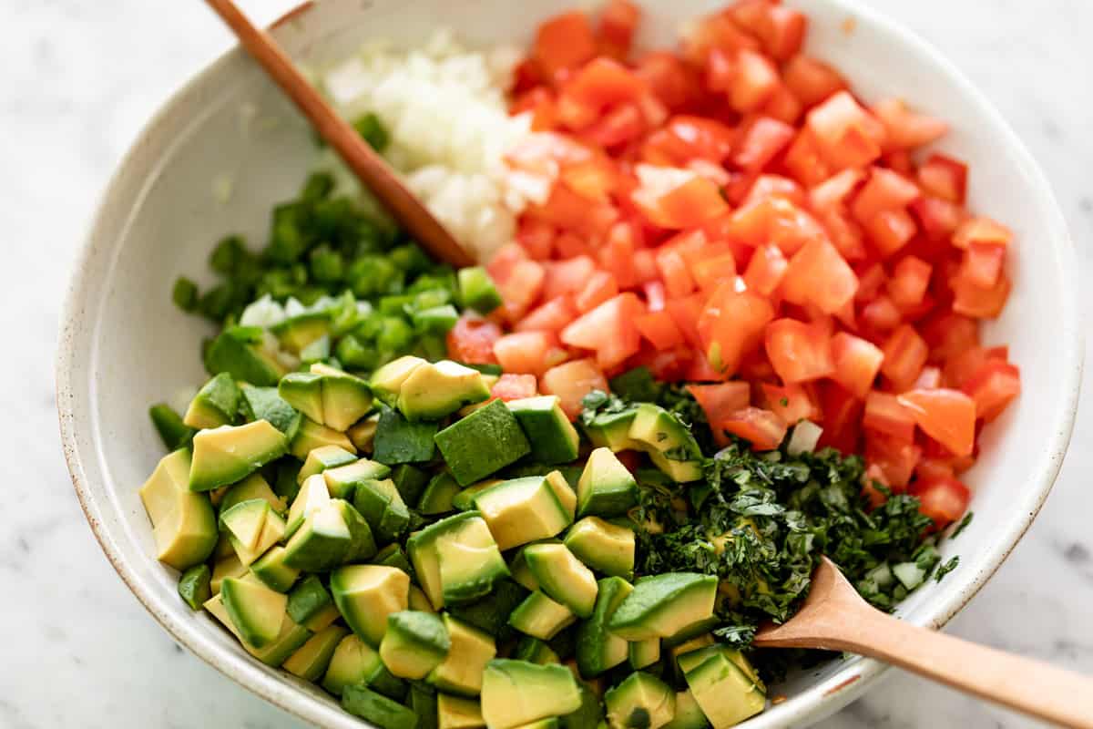 Avocado, tomato, capsicum, onion and herbs in a bowl with salad spoons. 