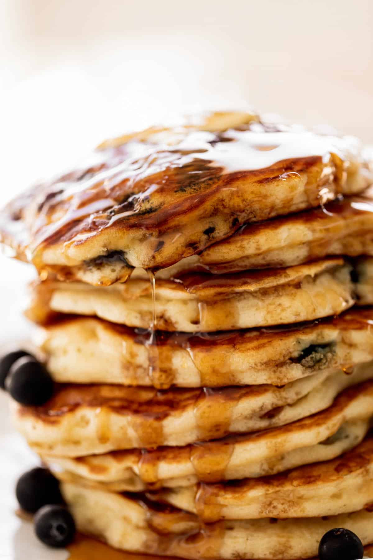 A stack of Blueberry Pancakes on a plate with maple syrup. Topped with fresh blueberries.