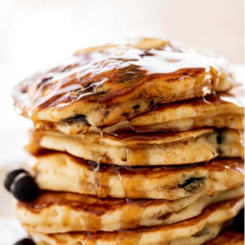 A stack of Blueberry Pancakes on a plate with maple syrup. Topped with fresh blueberries.