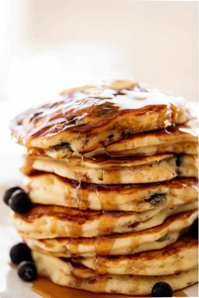 A stack of Blueberry Pancakes on a plate with maple syrup. Topped with fresh blueberries.