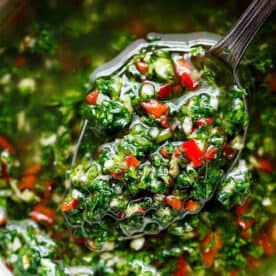 A small serving spoon lifts a spoonful of vibrant chimichurri, showcasing green herbs and red chili pieces mixed with oil from a bowl.
