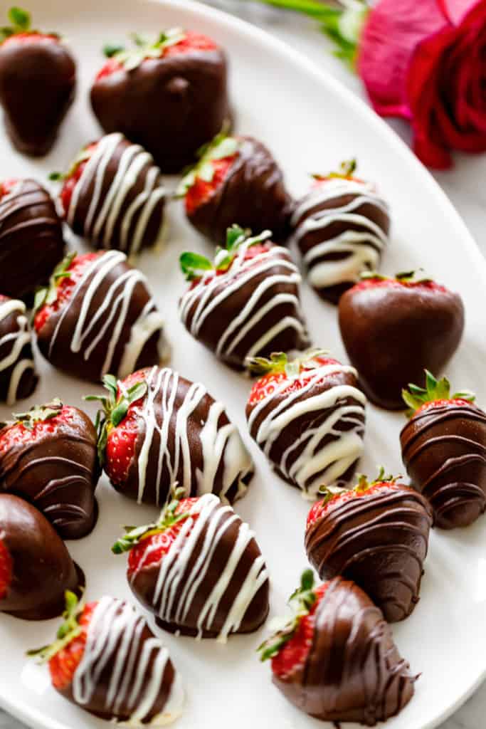 A white plate full of alcohol infused Chocolate Covered Strawberries. A rose sits behind the plate for romantic decoration. | cafedelites.com