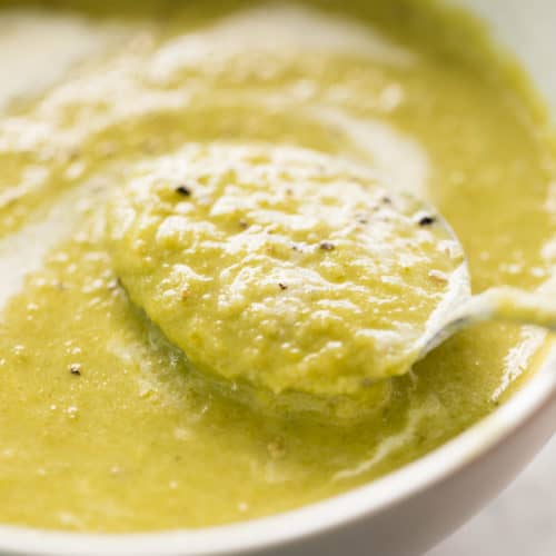 A comforting cream of asparagus soup garnished with cracked black pepper served in a white bowl with a silver metal spoon.