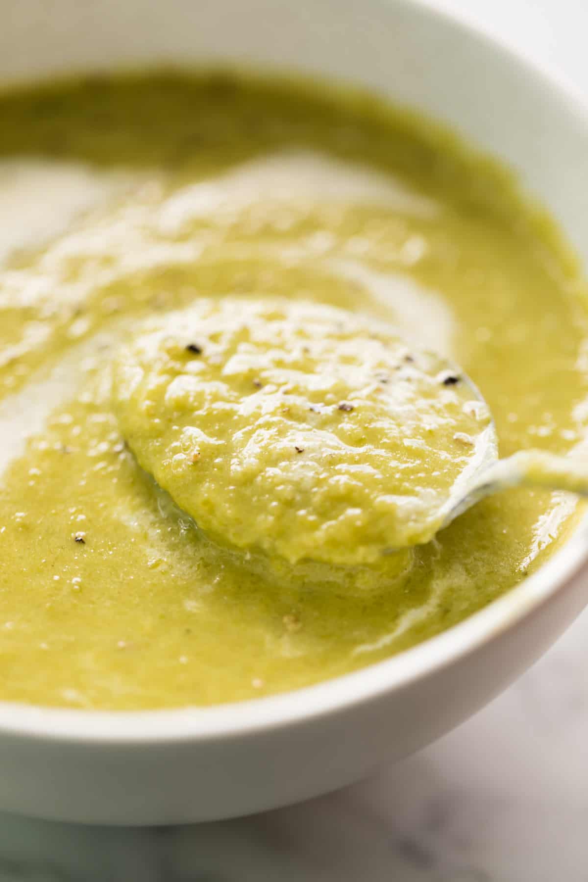 A comforting white bowl of cream of asparagus soup garnished with cracked black pepper. Served with a silver metal spoon in the soup ready to eat!