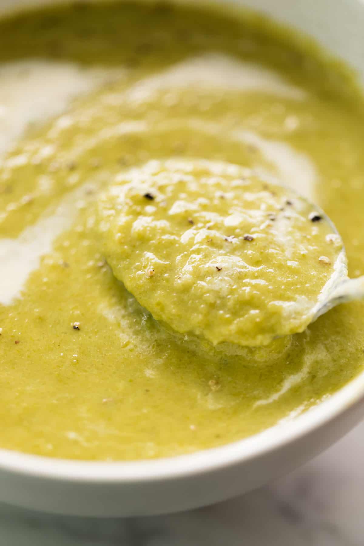 A comforting white bowl of cream of asparagus soup garnished with cracked black pepper. Served with a silver metal spoon in the soup ready to eat!