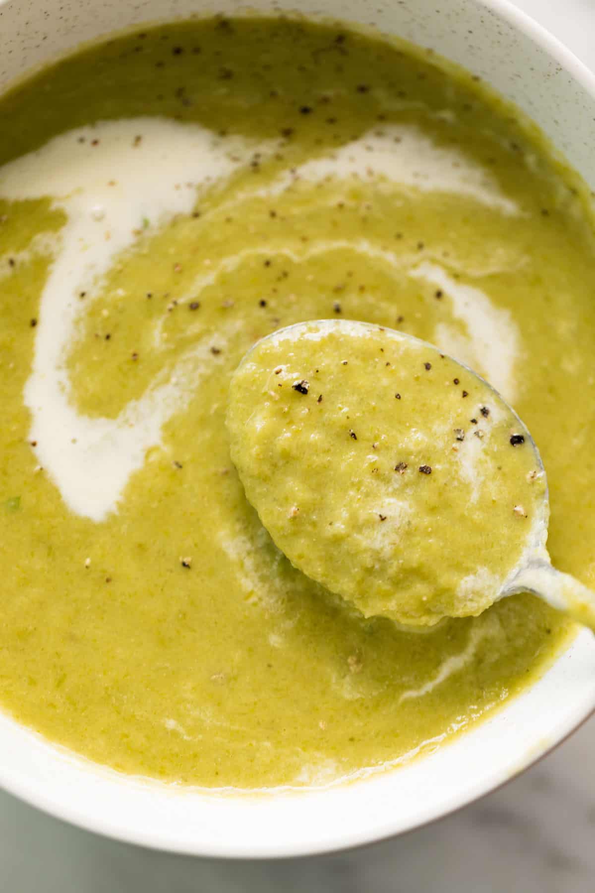 A comforting white bowl of cream of asparagus soup garnished with cracked black pepper. Served with a silver metal spoon in the soup ready to eat!