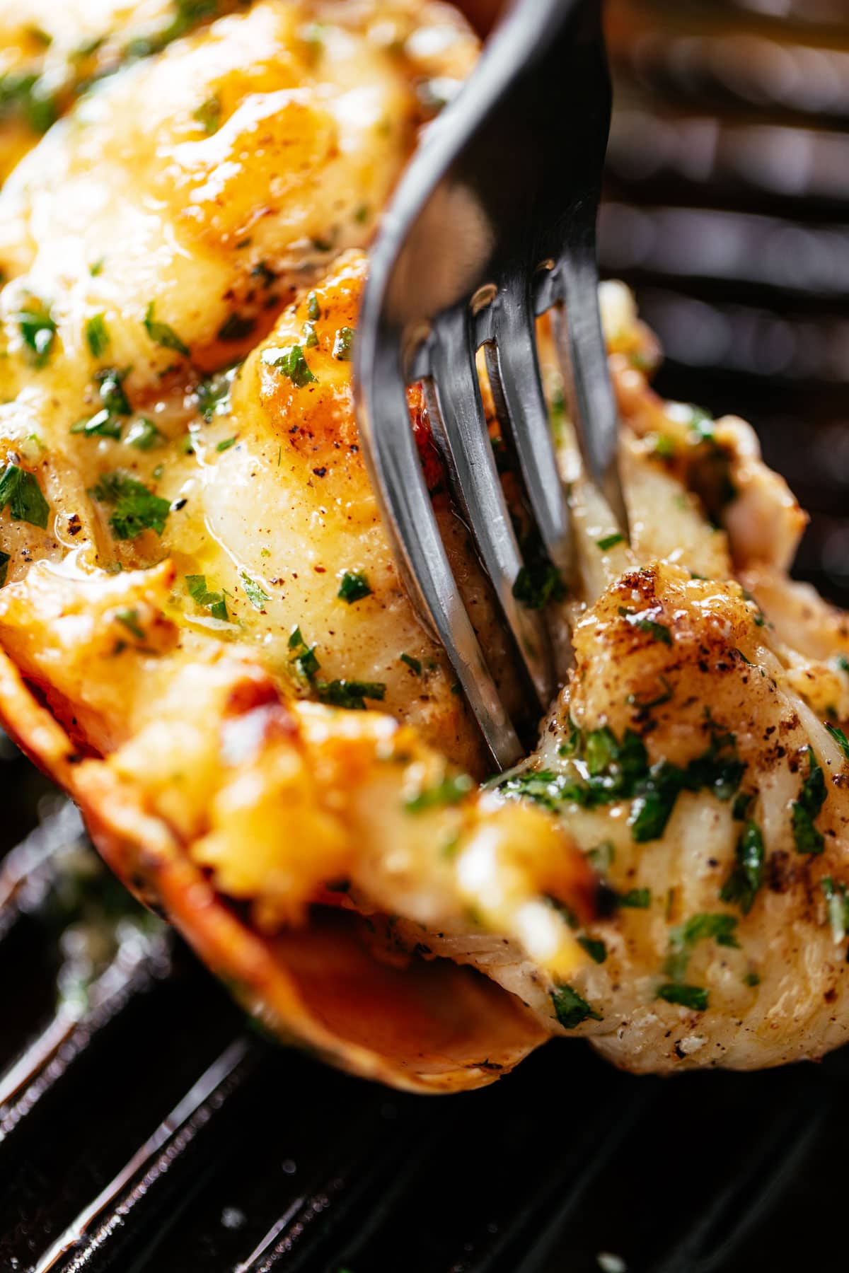 A fork pierces through a grilled lobster tail brushed with melted garlic herb butter on a black grill pan | cafedelites.com