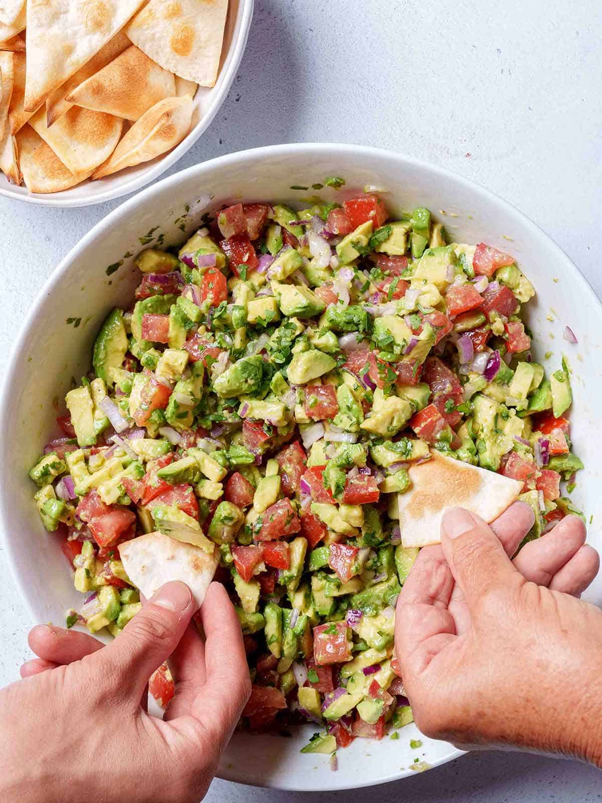 An image of Avocado Salsa Verde in a white bowl being scooped up with a tortilla chips by two hands.