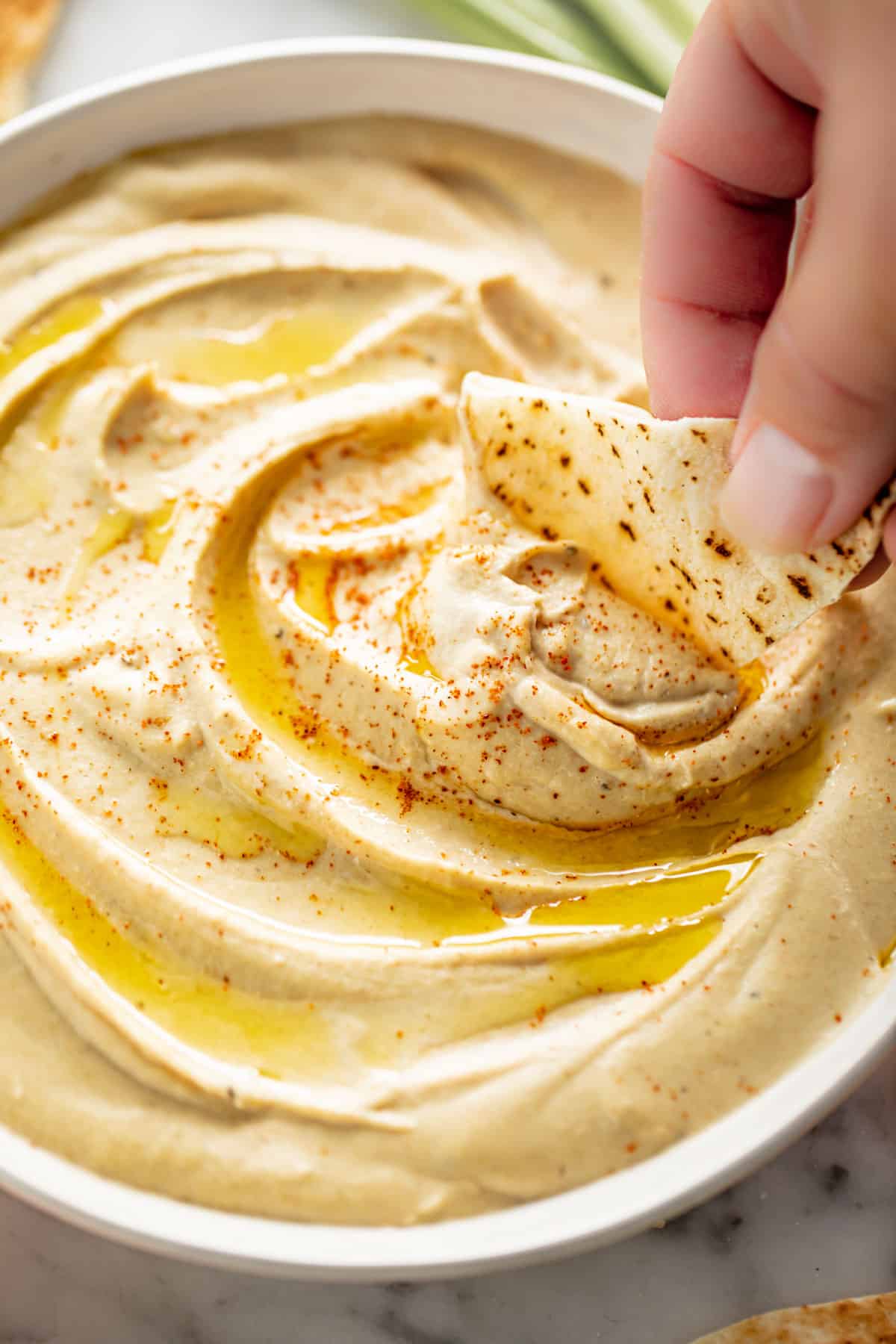 A side view image of a piece of pita bread dipped into Hummus served in a white bowl, drizzled with olive oil and topped with a sprinkle of paprika.