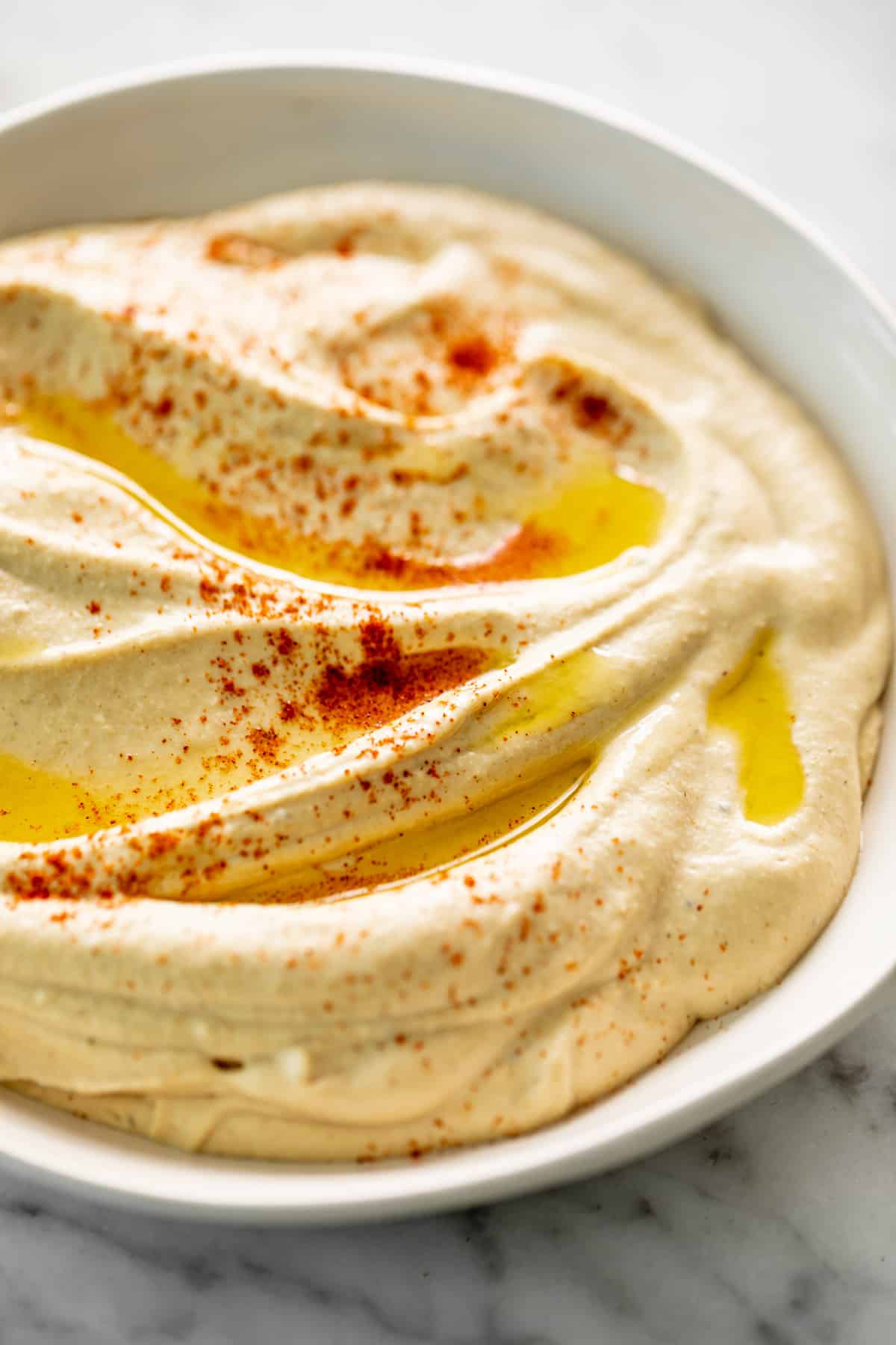 A side view image of Hummus served in a white bowl, drizzled with olive oil and topped with a sprinkle of paprika.