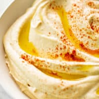 A side view image of Hummus served in a white bowl, drizzled with olive oil and topped with a sprinkle of paprika.