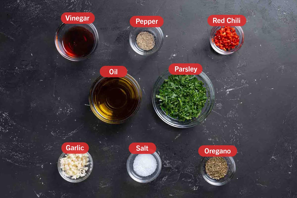 All the ingredients needed for the preparation of the chimichurri, specifically: vinegar, pepper, red chili, oil, parsley, garlic, salt, and oregano.