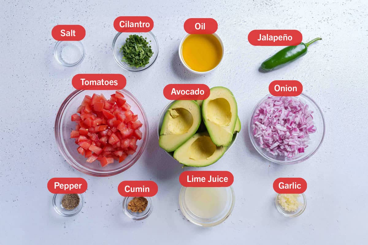 Image of all the ingredients needed to prepare the avocado salsa: avocado, tomatoes, onion, salt, cilantro, oil, jalapeño, pepper, cumin, lime juice, and garlic.