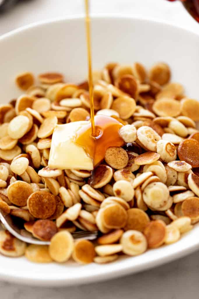 Mini Pancakes Cereal served with a piece of butter and drizzled with maple syrup. Served in a white bowl with a silver fork. | cafedelites.com