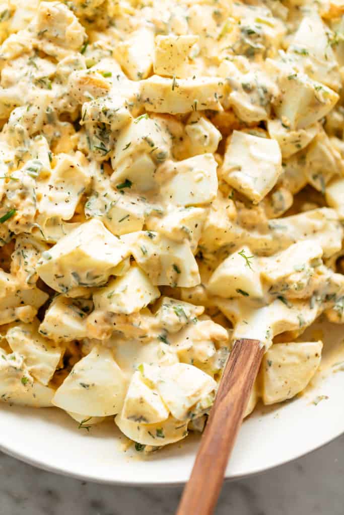 A white serving bowl full of the most perfect egg salad with a creamy mayo dressing. A small wooden bowl of fresh herbs to garnish sits in the background.