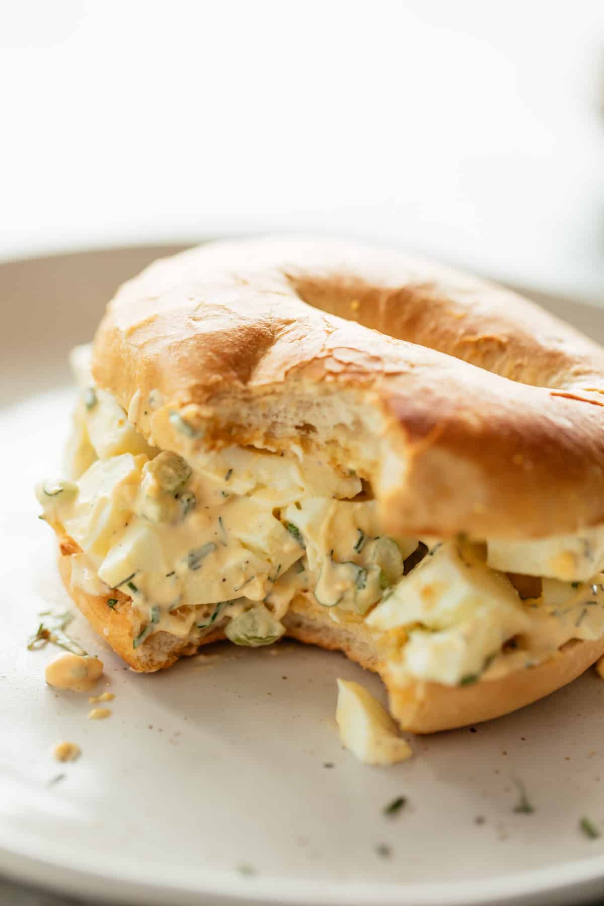 Perfect Egg Salad served sandwiched in a bagel on a white plate sprinkled with fresh dill, chives and parsley. | cafedelites.com