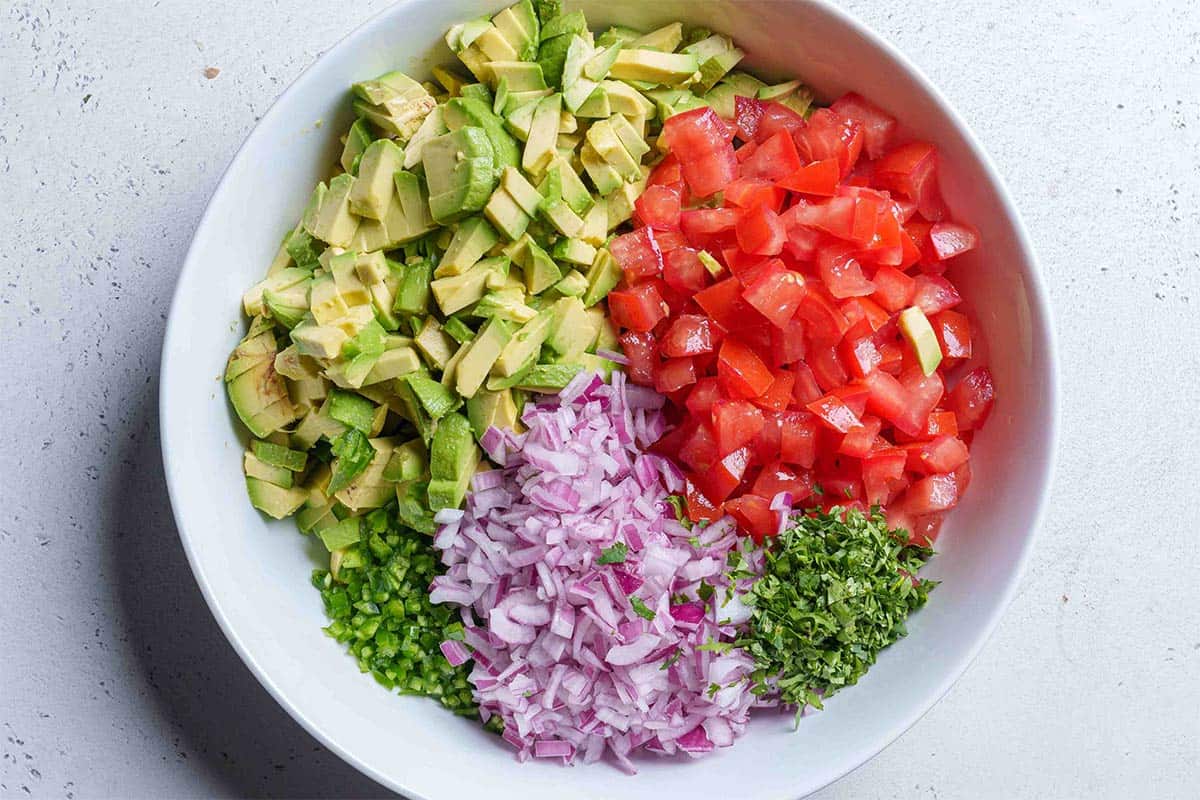 Step 2: The image depicts finely chopped chunks of avocado, tomatoes, onions, cilantro, and jalapeños in a white mixing bowl.