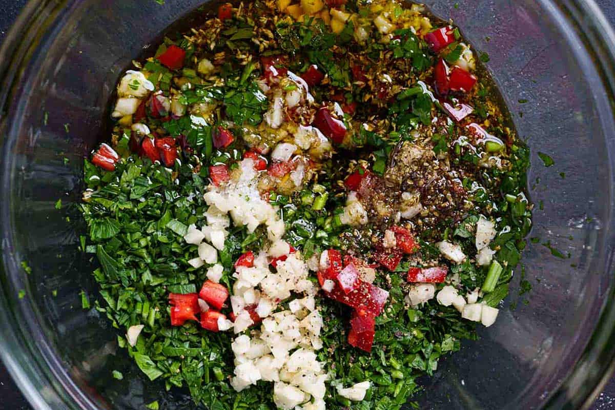 The image shows olive oil, red wine, vinegar, and oregano being added to the mixture of previously chopped ingredients.