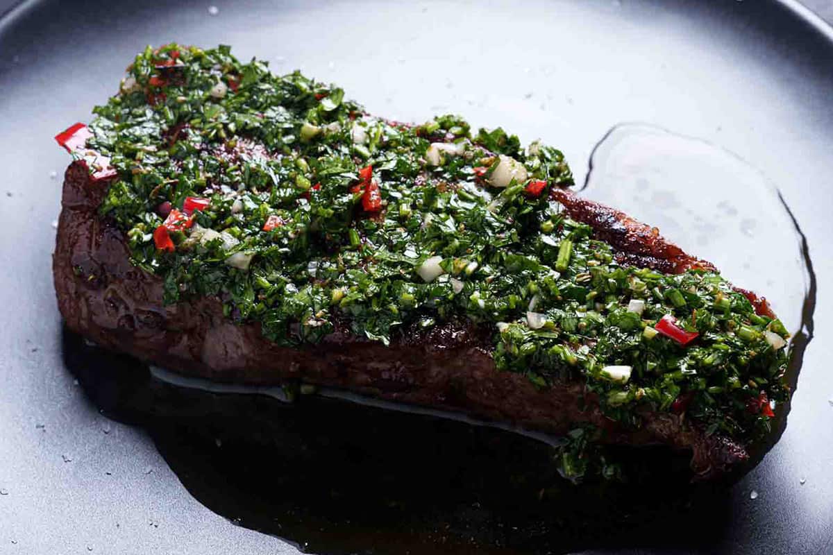 An image of a beautiful rump steak topped with finely chopped chimichurri, with oil pouring from it.