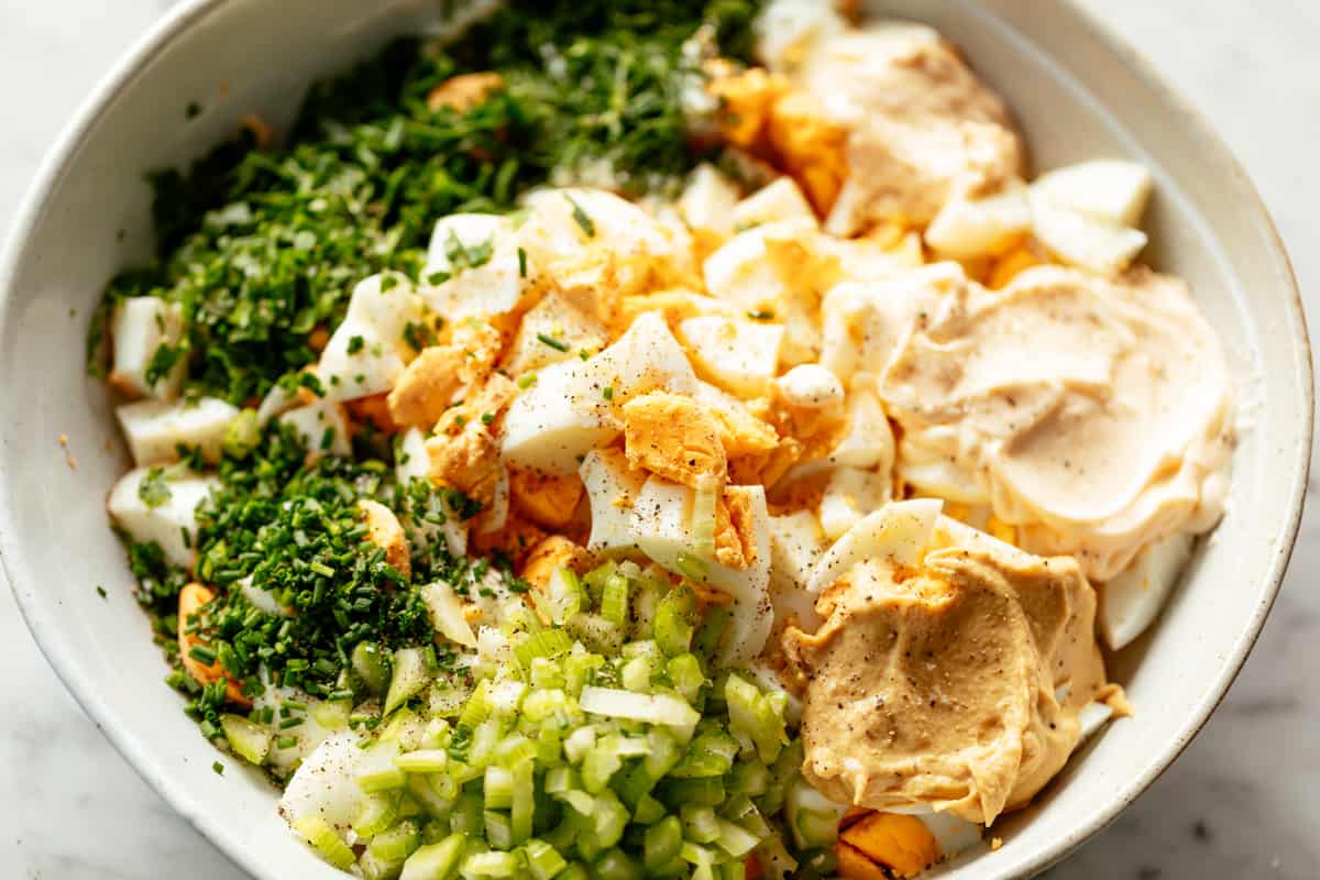 A white serving bowl full of ingredients shown for egg salad recipe. Include chopped eggs in the centre surrounded by (clockwise from top right) cream of horseradish, mayonnaise, Dijon mustard, finely chopped celery, fresh chopped chives, fresh chopped dill, fresh chopped parsley. All seasoned with salt and pepper. | cafedelites.com