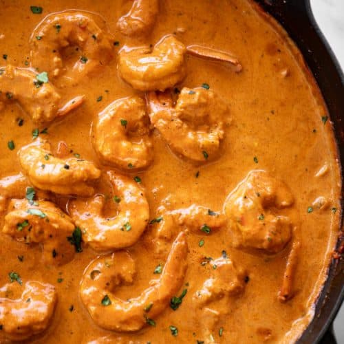 A black cast iron skillet, labeled "Staub," is filled with a rich, orange-red shrimp curry. The curry is garnished with fresh, green cilantro and is teeming with plump, cooked shrimp. A silver serving spoon rests in the curry, ready to enjoy.