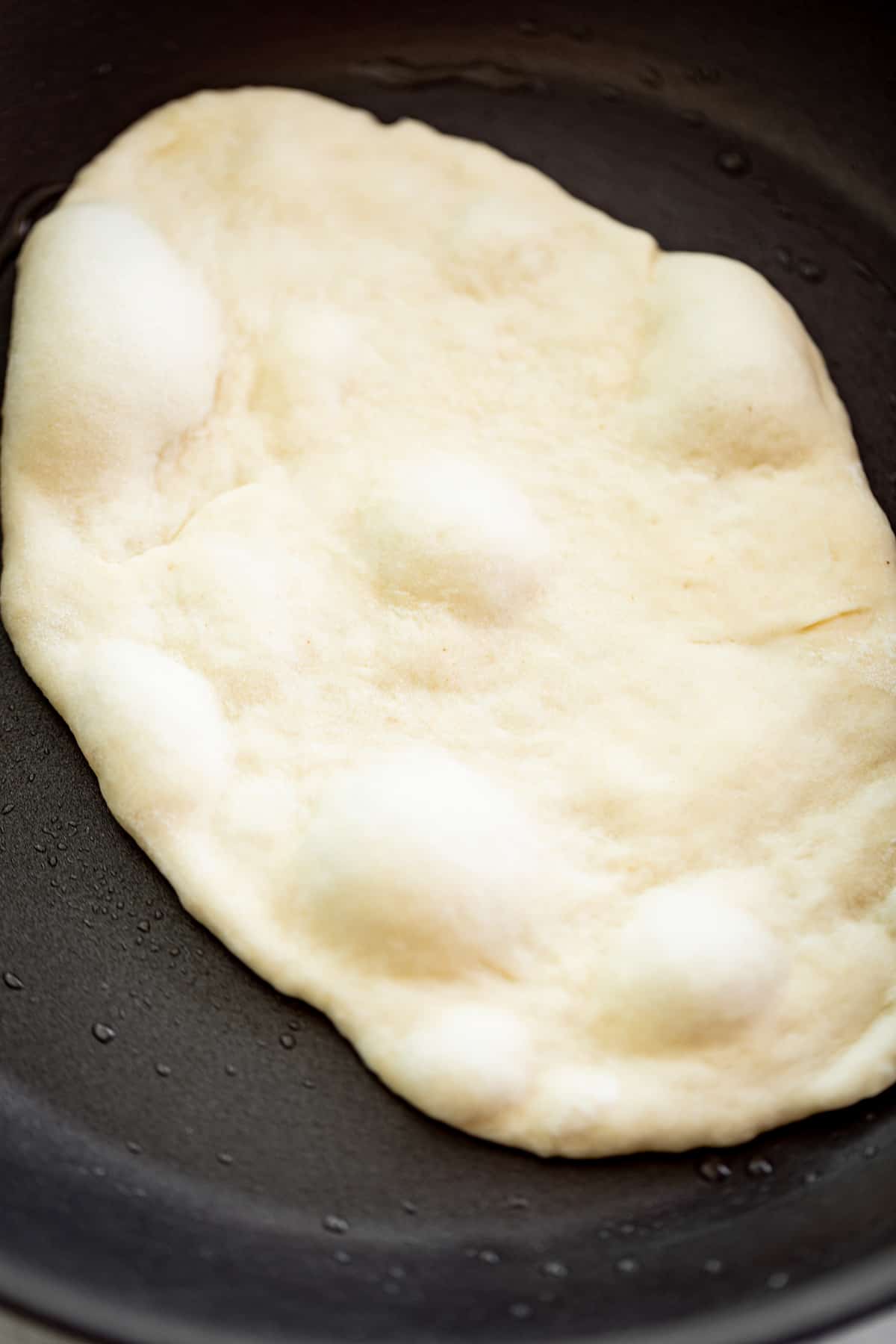 An image of Cooking a garlic naan bread in a black skillet with a light coating of oil.