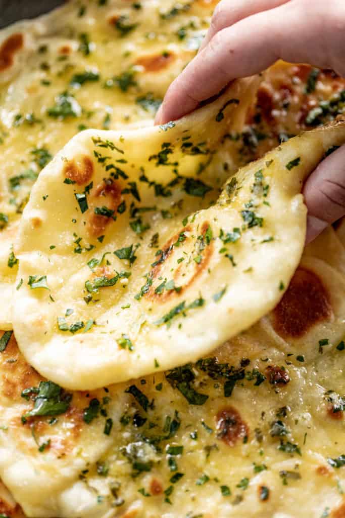 Holding a Garlic Naan Bread on a plate with a pile of naan. Garnished with fresh chopped cilantro. | cafedelites.com