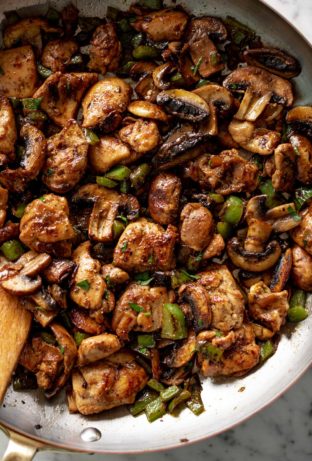 A wooden spoon stirring a pan filled with cooked mushrooms, chopped green onion and thick chicken pieces.