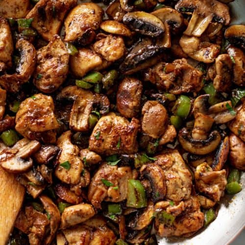 A wooden spoon stirring a pan filled with cooked mushrooms, chopped green onion and thick chicken pieces.